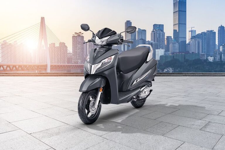 Gray scooter parked on a wide paved plaza with a city skyline and a bridge in the background, at sunset.