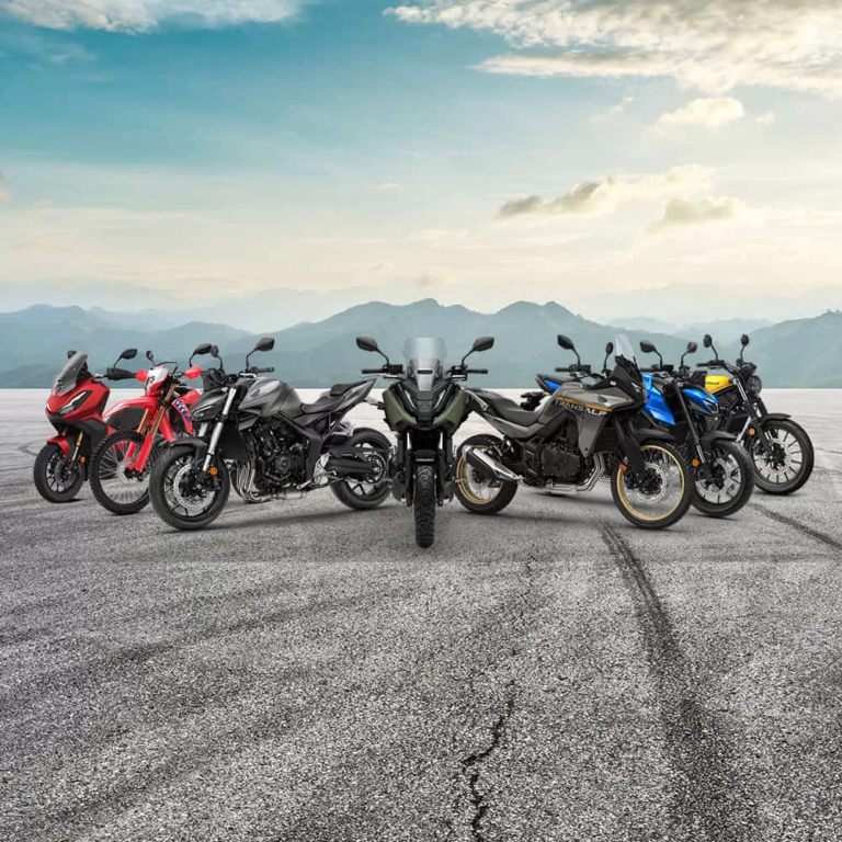 Seven motorcycles parked in a row on a cracked tarmac with distant mountains and blue sky in the background, forming a lineup.